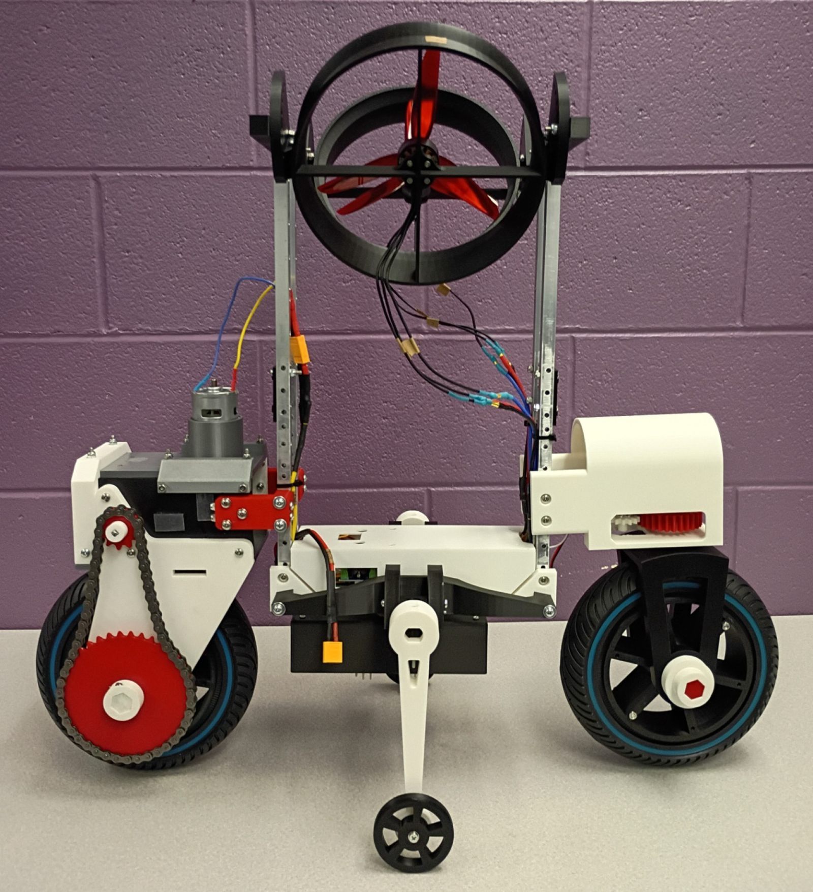 The finished bike on a table: two ducts on a mast above a white 3D-printed body, red chain drive at the rear wheel, a small side-support wheel below